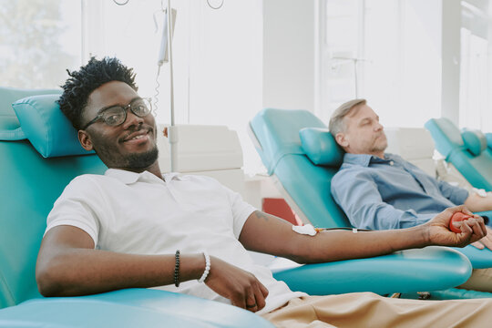 Young Black man donating blood while sitting in medical chair, smiling at camera, middle aged Caucasian man in background also donating blood, both participating in blood donation process - Powered by Adobe