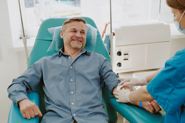 Middle aged Caucasian man sitting in medical chair donating blood, while female healthcare worker...
