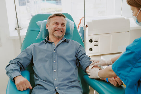 Middle aged Caucasian man sitting in medical chair donating blood, while female healthcare worker preparing arm for blood draw in clinical setting with medical equipment visible - Powered by Adobe