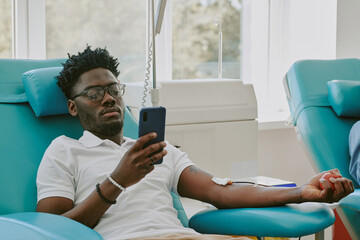 Young adult Black man donating blood while reclining in medical chair, holding smartphone in one hand, medical tubing attached to arm, participating in blood donation process at clinic