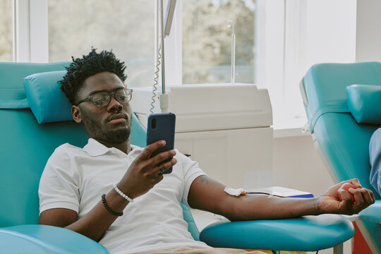 Young adult Black man donating blood while reclining in medical chair, holding smartphone in one hand, medical tubing attached to arm, participating in blood donation process at clinic