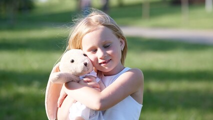 A girl with long blonde hair hugs a soft bunny toy while standing in a bright, green park, surrounded by trees and enjoying a sunny afternoon.