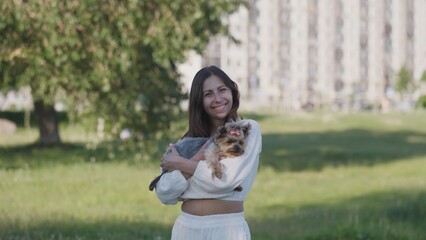 Bright sunlight fills the park as a woman smiles while holding her small dog, creating a cheerful atmosphere surrounded by nature and urban buildings.
