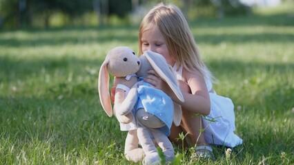A cheerful girl interacts with her soft bunny toy, sharing a joyful moment on a bright, sunny day surrounded by green grass in the park.