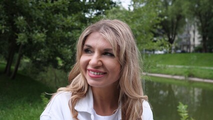 A woman with long blonde hair smiles warmly while seated near a calm body of water in a lush park. Sunlight filters through the trees creating a serene atmosphere.
