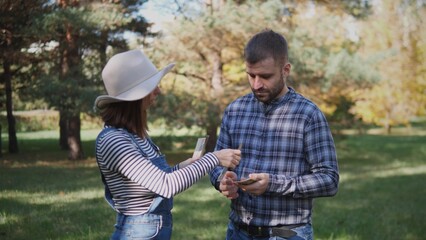 Two friends stand in a green park exchanging plants while enjoying a sunny day. The bright foliage creates a vibrant atmosphere for their conversation.