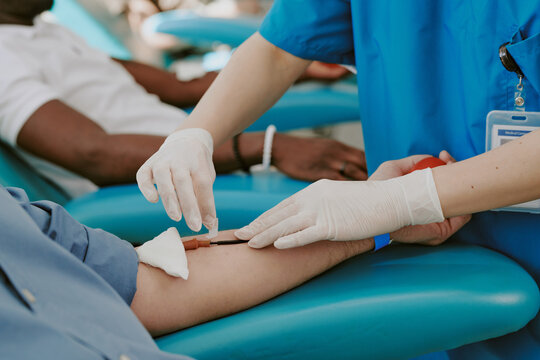 Caucasian middle aged man donating blood while medical worker wearing gloves inserting needle into arm, Black man visible in background also participating in blood donation process