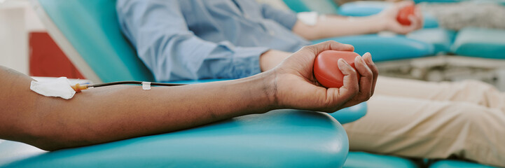 Black young adult man donating blood in medical facility, holding stress ball in hand while blood collection tube attached to arm, Caucasian donating blood in background