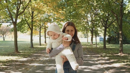 A cheerful woman carries a young child dressed warmly in a soft outfit as they walk among golden leaves in a peaceful park during the fall season.
