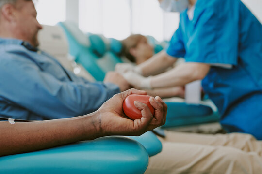 Black young adult man squeezing stress ball while donating blood in medical clinic, Caucasian middle aged man and woman receiving blood donation procedure in background, nurse assisting