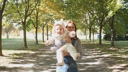 A woman carries a cheerful child dressed in a white suit with bear features, both laughing in a park filled with colorful autumn foliage on a bright day.