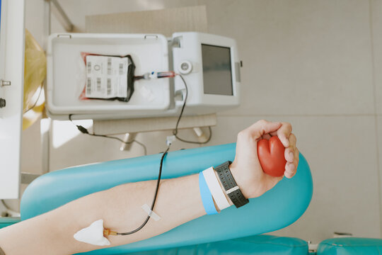 Caucasian young adult man donating blood, arm extended with needle inserted, holding red stress ball, blood collection bag and medical device visible on table during donation process