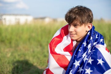 Happy teenage boy with American flag on shoulders in field outside city