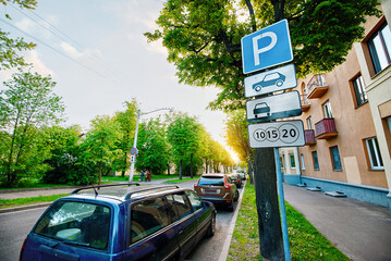 Parked cars under leafy trees in residential area, paid parking zone sign on pole, issue of limited free parking spaces. Roadside view in green residential zone with vehicles parked closely under tree