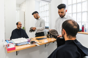 Hairdressers showing different hairpieces to a balding customer sitting in front of a mirror in a...
