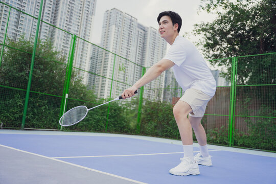 Young man practices badminton on outdoor court in urban area during cloudy day while focusing on perfecting technique and form