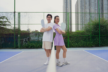 Couple poses proudly with pickleball paddles on an urban court surrounded by high-rise buildings in...