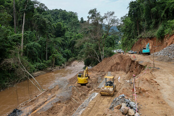 The landscape of the rural road is being repaired by a backhoe by a road contractor. The road cuts into the forest along the river from a landslide during the rainy season.
