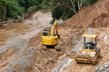The landscape of the rural road is being repaired by a backhoe by a road contractor. The road cuts into the forest along the river from a landslide during the rainy season.