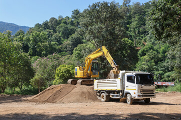 A white truck is waiting for a yellow backhoe to load sand into the back of a construction site surrounded by forest.
