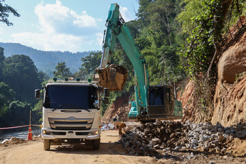 A white truck is waiting for a green backhoe to load sand into the back of a construction site surrounded by forest.