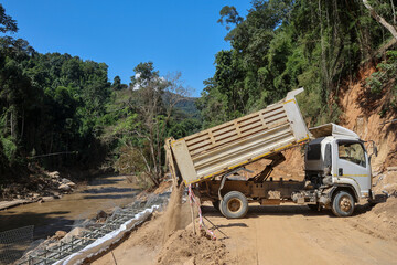 A white truck is dumping sand on the ground on a rural road through a forest being repaired, in the construction area of ​​a road repair contractor.