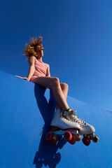 Young woman wearing roller skates sitting on blue wall under vivid blue sky