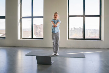 A young girl is practicing yoga indoors, following online instructions displayed on a laptop,...