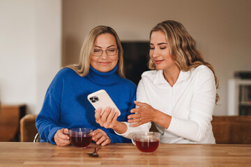 Women sharing a moment over tea while enjoying a conversation and looking at a smartphone in a cozy indoor setting