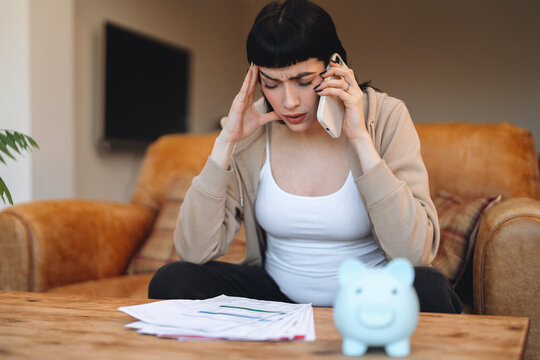 Woman sitting on a couch with a concerned expression while talking on the phone about finances in a cozy living room - Powered by Adobe