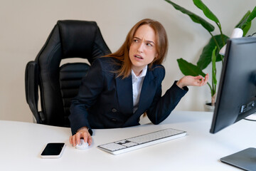 Professional woman expressing confusion while working at a modern desk with computer and smartphone...