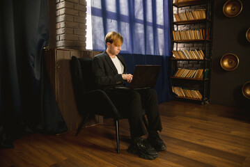 Young man with red hair sits in a chair, working on a laptop in a room with a bookshelf and window....