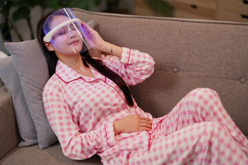 Young Asian adult woman unwinding on a comfortable sofa utilizing an innovative LED light therapy...