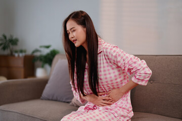 Young Asian adult woman suffering from severe abdominal pain or period cramps at home on sofa...