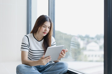 Beautiful young asian woman is using a tablet while sitting on the floor next to a large window in a modern office building, looking focused and productive