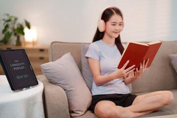 Adult Asian woman enjoying longevity podcast on tablet and reading a book with headphones for...