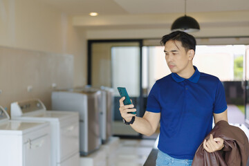 Young Asian man using smartphone in laundry room managing washing with mobile app, checking time, making online payment or video chatting with family