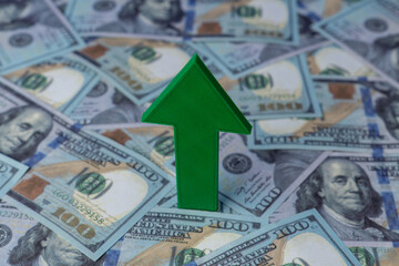 A green arrow points upwards against a background of scattered US one-hundred dollar bills. Financial growth, economic prosperity, currency appreciation concept.