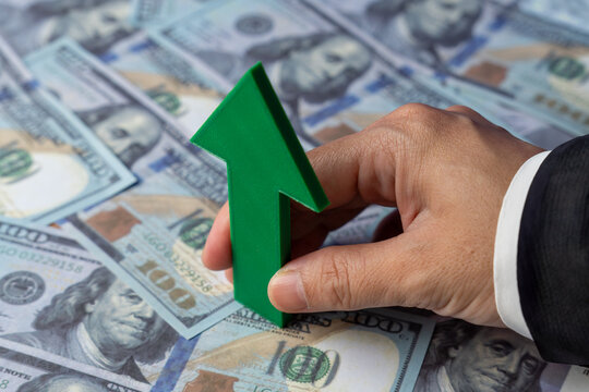 A hand holds a green upward-pointing arrow over a spread of US one-hundred dollar bills. Currency appreciation and strength, investment gains, recovering economy concept.