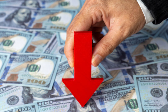 A hand holds a red arrow pointing downwards against a background of scattered US one-hundred dollar bills. Financial downturn, economic loss, declining markets, and currency depreciation concept.