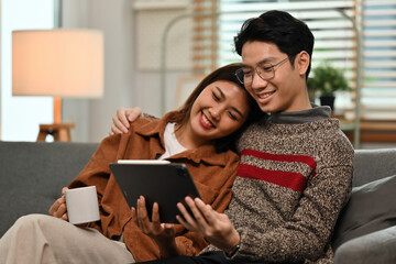 Happy couple sitting close on a couch, sharing a tablet and coffee in a cozy home