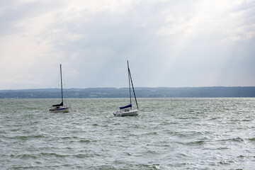 Two yachts on a wavy lake under a grey sky. Light beams shine through the clouds, adding contrast...