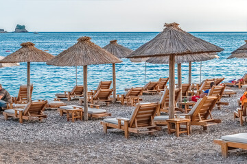 Plaža Župlica in Budva, Montenegro, with thatched umbrellas, wooden lounge chairs on a pebble beach, and calm Adriatic waters in the background as people relax by the shore