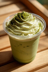 Creamy whipped matcha latte in plastic cup topped with green tea powder, placed on wooden surface with warm sunlight