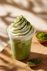 Creamy whipped matcha latte in plastic cup with green tea powder on wooden table under warm sunlight