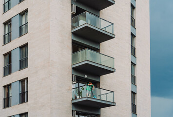 Woman cleaning a glass balcony with a mop and spray bottle, seen from a neighboring building. Urban...