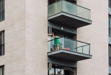 Woman cleaning a glass balcony with a mop and spray bottle, seen from a neighboring building. Urban...