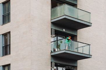 Woman cleaning a glass balcony with a mop and spray bottle, seen from a neighboring building. Urban residential facade with multiple balconies and modern architectural elements