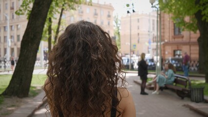 In a bustling city park, a person with curly hair strolls past benches and passersby, soaking in the warm sun and lively ambiance of the urban landscape.