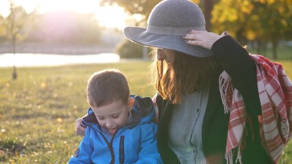 A woman wearing a hat sits beside her young son, both smiling as they share a playful moment on the grass in a park during a sunny autumn afternoon.